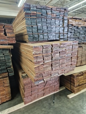 Large stacks of cut timber planks with black-coated ends stored in a warehouse under fluorescent lighting