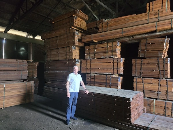 Man standing in front of large stacks of processed timber planks inside a dimly lit warehouse